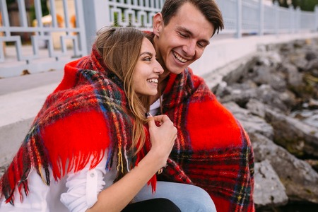 beautiful young couple sitting on a stone embankment, wrapped in a blanketの写真素材