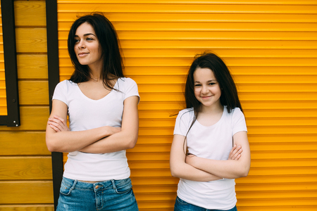 Beautiful young mother and her cute little daughter, looking at camera and smiling, against wallの写真素材