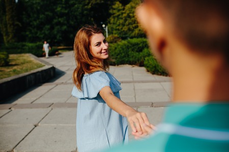 Young woman leading her lover by hand in park, looking at him, playfully laughingの写真素材