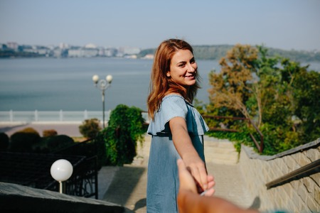 Young woman leading her lover by hand in park, looking at him, playfully laughingの写真素材