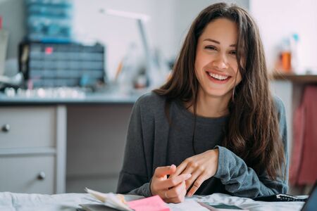 Portrait of smiling cute hipster girl staring on camera while writing college exercises in notebook.の写真素材
