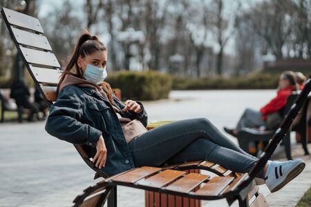 Yong woman sitting on the bench in the park and wearing a protective mask. Coronavirus concept.の写真素材