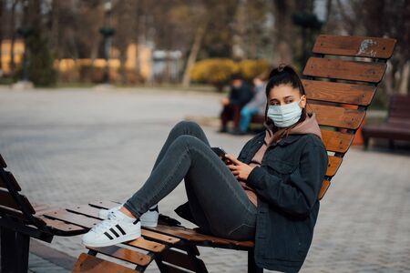 Yong woman sitting on the bench in the park and wearing a protective mask. Coronavirus concept.の写真素材