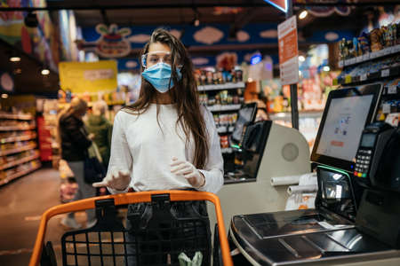 A young woman scans products in a mask and gloves. Self-checkout counter in the food store during coronavirus epidemic.のeditorial素材