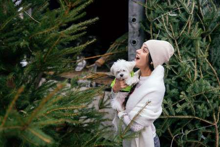 Woman with a white dog in her arms near a green Christmas trees