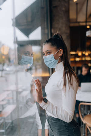 Young woman in face mask standing in front of windows in cafe.の写真素材