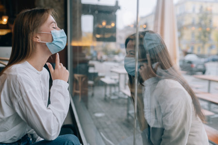 Two women in protective masks opposite each other, window between them.の写真素材