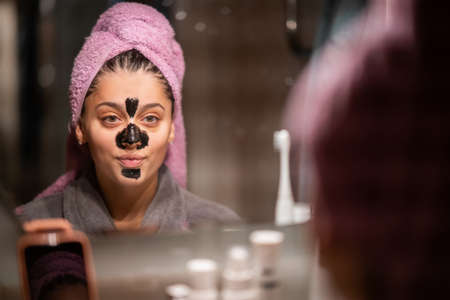 Beautiful woman with a clay or a mud mask on her face looking in the mirror