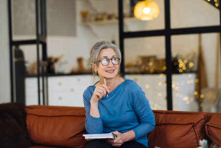 Happy senior woman in glasses thinking and writes in a diary homeの写真素材