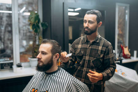 Cheerful young bearded man getting haircut by hairdresser at barbershopの写真素材