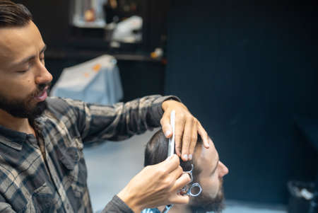 Cheerful young bearded man getting haircut by hairdresser at barbershopの写真素材