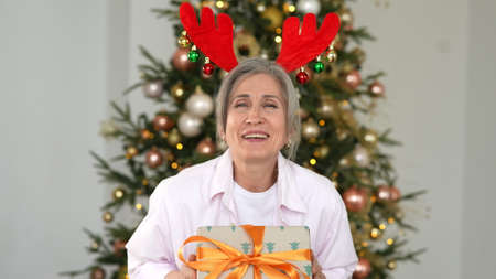 Grandma wearing deer christmas hat. Christmas presents in hand talking at cameraの写真素材