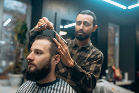 Cheerful young bearded man getting haircut by hairdresser at barbershopの写真素材