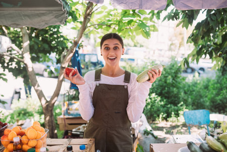 Young saleswoman holding zucchini and tomato in handsの写真素材