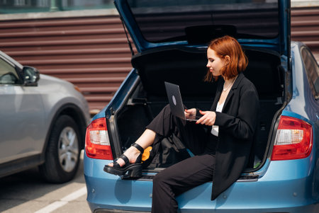 Woman working on laptop while sitting in trunk of carの写真素材