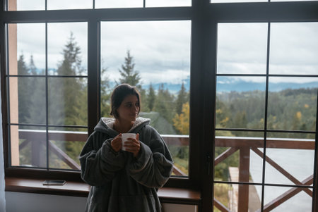 Young woman standing by the window holds a mug