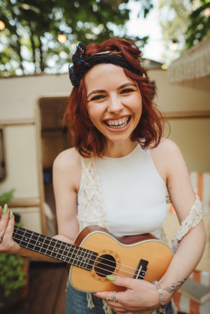 Happy hippie girl is having a good time with playing on guitar in camper trailer. Holiday, vacation,...