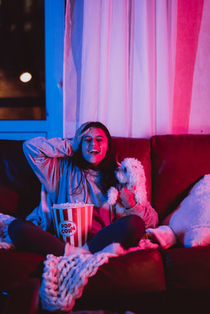 Beautiful young girl watching movie at home in a dark room with a popcorn bucket and playing with her dog and sits in front of a monitor or TV.の写真素材