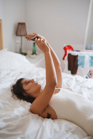 In a cozy bedroom, a young woman happily uses her smartphone to connect with othersの写真素材