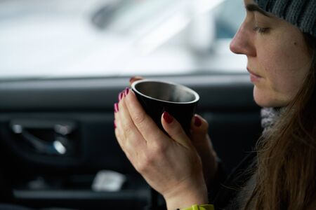 Young woman sits in the trunk of the car and holds a cup of hot drink in her handsの写真素材