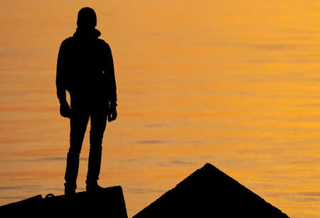 Woman or man standing on rock looking straight. Nature and beauty concept. Orange sundown. Girl silhouette at sunsetの写真素材