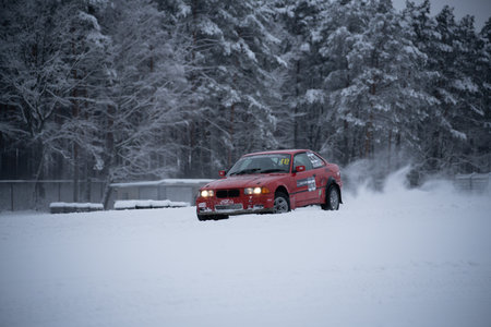 12-12-2022 Riga, Latvia a red car driving through a snow covered forest area with trees in the background and a dusting of snow on the ground. .のeditorial素材