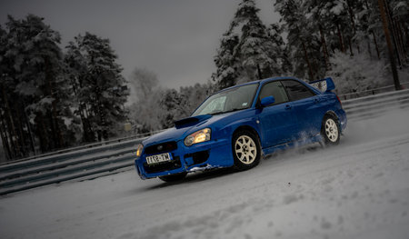 12-12-2022 Riga, Latvia a blue car driving down a snowy road next to trees and a fence with snow on it's sides. .のeditorial素材