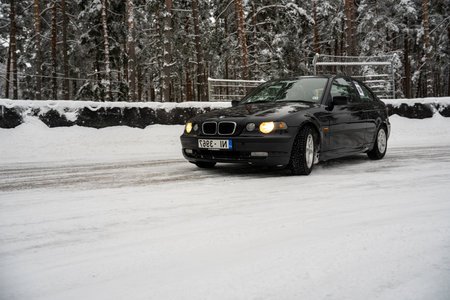 12-12-2022 Riga, Latvia a black car driving down a snowy road next to a forest of trees and a fence with a snow covered fence. .のeditorial素材
