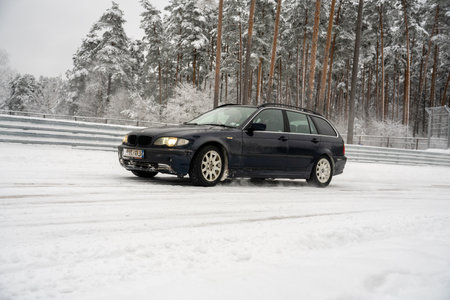 12-12-2022 Riga, Latvia a black car driving down a snow covered road next to a forest of trees and a fence with snow on it. .のeditorial素材