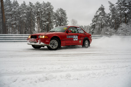 12-12-2022 Riga, Latvia a red car driving down a snow covered road next to a forest of trees and a fence with snow on it. .のeditorial素材