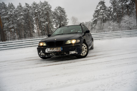 12-12-2022 Riga, Latvia a black car driving down a snowy road next to a forest of trees and a fence with snow on it. .のeditorial素材