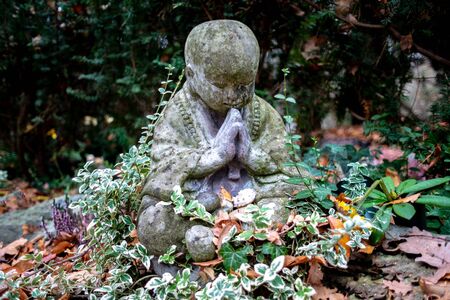 Stone statue of a little weeping buddha sitting on a grave sourrounded by plantsの写真素材