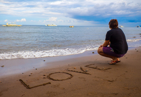Inscription Love on the sand. Empty beach with a man in black summer clothes. Romantic landscape with sea and lonely person. Love word near waterline. Tropical island vacation. Holiday romance photoの写真素材