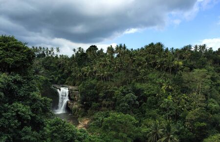 Huge waterfall in tropical forest. Bright exotic nature with fresh water stream. Dark green trees and white waterfall under cloudy sky. Distant lake and mountain in green bush. Travel in Bali islandの写真素材