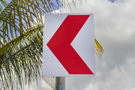 Red arrow pointer on tropical background. Roadside with palm tree leaves and cloudy sky. Road turn sign. Summer holiday travel concept. Serpentine way attention sign. Highway turn. Safe drive imageの写真素材