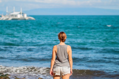 Woman in shirt and shorts on beach. Bright photo of young girl watching cruise boat. Seaside vacation travel vintage banner template. Turquoise water of tropical lagoon. Sunny day by sea backgroundの写真素材