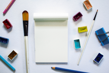 Art tools and blank notepad on white table. Painting art supply - brush, watercolor, pencil. Artistic flat lay on white table top view. Creative hobby or handmade craft banner template with text placeの写真素材