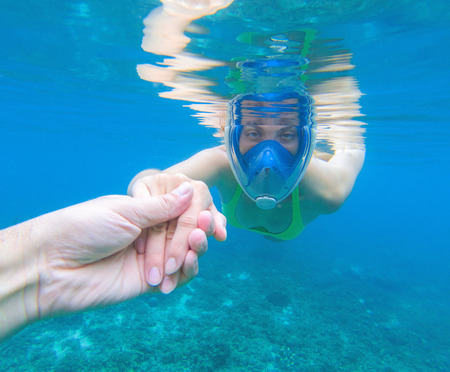 Snorkeling woman in full-face snorkeling mask. Swimming girl holds hand of partner. Romantic underwater photo. Snorkeling lesson. Sea water sport. Coral reef snorkeling. Swimming woman guiding by handの写真素材