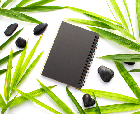 Bamboo leaf and black paper background. Black paper notebook mockup. Green bamboo leaf and beach pebbles flat lay. Blank notepad top view. Spa beauty banner template with text place. Bamboo decorの写真素材