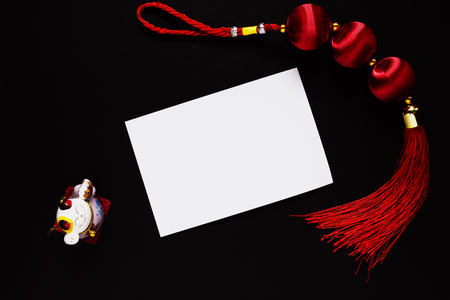 Chinese lucky knot, maneki neko cat figurine and empty paper card on black. Chinese New Year decor. White paper card. Chinese Lunar New Year top view photo. Asian winter holiday greeting card flat layの写真素材