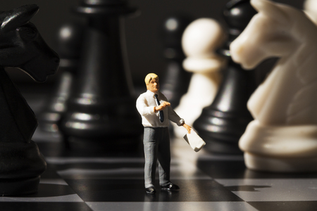 Miniature teacher explains chess game rules on board. Chess strategy analysis concept. Black and white chess figurines and businessman. Learning chess rules. Chessboard game play process macro photoの写真素材