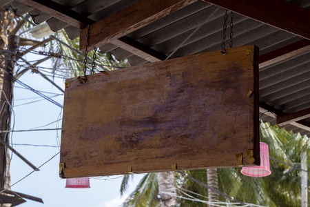 Blank wooden board sign hang under roof. Tropical island cafe or hotel sign mockup. Wooden board with palm leaf background. Exotic place for vacation. Empty billboard outdoor. Rustic board textureの写真素材