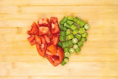 Tomato and okra vegetables in heart shape on cutting board. Vegetables cut top view photo on wooden background. Healthy vegetarian salad preparation. Fresh summer food. Slimming diet. Raw eatingの写真素材