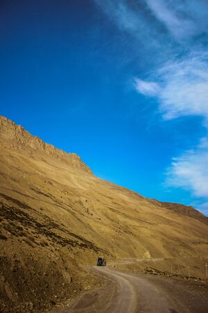 Yellow-Brown Desert Mountain in Spiti Valley, Himachal Pradesh, India. Against the plain blue sky, with a tractor going in the distance. Serene martian-looking background landscape.の写真素材