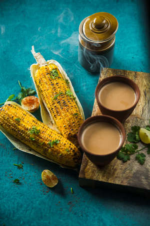 Top view of brewed hot milk tea with roasted grilled corn on blue background. Delicious Grilled Corncobs and Two Cups of Chai, an ideal Indian monsoon brunch good for boosting immunity and health.の写真素材