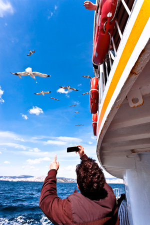 Feeding flying seagulls, ferriboat, people, blue sea, waves, bright sky, small white cloudsの写真素材
