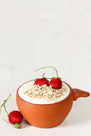 oat flakes with sour cream and strawberry in a clay pot on a white wooden tableの写真素材