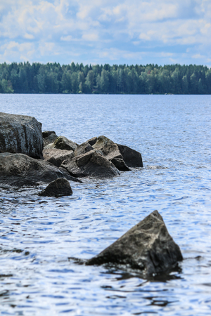 Huge calm blue summer lake with green taiga forest in background and big solid rocks on frontの写真素材