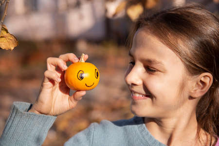 Smiling girl looks at a smal pumpkin with a smiley. Concept of joy and optimism.の写真素材