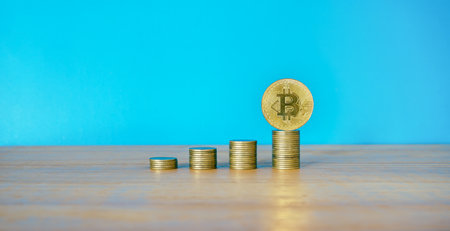 Golden bitcoin coin and stack of coins on wooden table with blue background.の写真素材